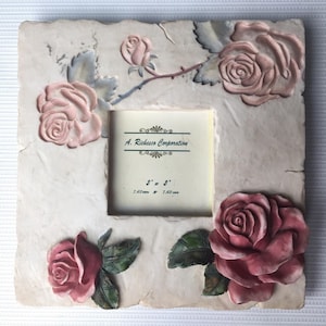 May include: Square photo frame with embossed pink roses and green leaves. The frame is off-white with a textured surface. The frame opening is 3" x 3" (7.62 cm x 7.62 cm). The frame has the text "A. Richesco Corporation".