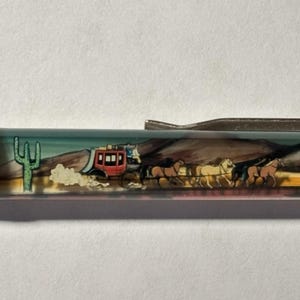 May include: A pen with a detailed illustration of a stagecoach pulled by horses, set against a desert landscape with a cactus. The pen's design features a red stagecoach and a mountain range.