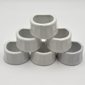 May include: A set of six white ceramic napkin rings arranged in a pyramid stack. Each ring features a smooth, glossy finish with a slightly rounded, square-like shape and vertical indentations along the outer surface, creating a uniform appearance.