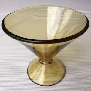 May include: A clear, amber-colored glass martini glass with a wide, shallow bowl and a dark brown rim. The stem is conical, leading to a circular base. The glass reflects light, highlighting its smooth surface.