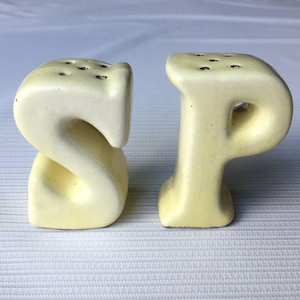 May include: A pair of vintage salt and pepper shakers. The shakers are shaped like the letters "S" and "P" in a pale yellow color. Each shaker has several holes on top for dispensing the contents. The set is made of ceramic.