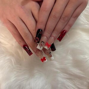 May include: Long, square acrylic nails with diverse designs. Some nails are solid red or black, while others feature Betty Boop designs and red bow accents. The nails are set against a fluffy, white background.