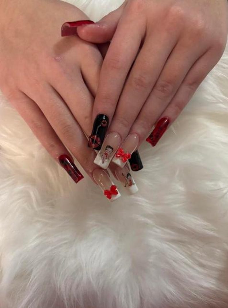 May include: Long, square acrylic nails with diverse designs. Some nails are solid red or black, while others feature Betty Boop designs and red bow accents. The nails are set against a fluffy, white background.