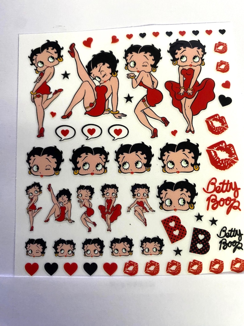 May include: A sheet of Betty Boop stickers. The stickers feature the cartoon character in various poses, all wearing a red dress. There are also hearts, stars, and the words "Betty Boop" in a cursive font. The background is white.