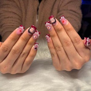 May include: Manicured nails with a light pink base, dark red tips, and white pearl accents. Each nail features a cartoon character, a heart, or a small design. The nails are square-shaped and have a glossy finish.