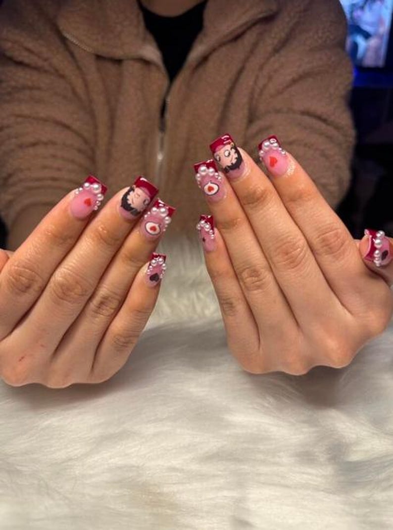 May include: Manicured nails with a light pink base, dark red tips, and white pearl accents. Each nail features a cartoon character, a heart, or a small design. The nails are square-shaped and have a glossy finish.
