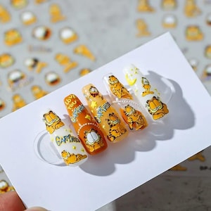 May include: A set of five artificial nails with Garfield designs. Each nail features a different Garfield image, with the word "Garfield" and the phrase "Love You" on some nails. The nails are orange, white, and yellow.