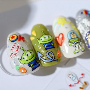 May include: Close-up of five fingernails with different designs. The designs include a green alien, a blue arrow, a heart, a cartoon astronaut, and a silver glitter design. The text "my best friend!" is written on one of the nails.