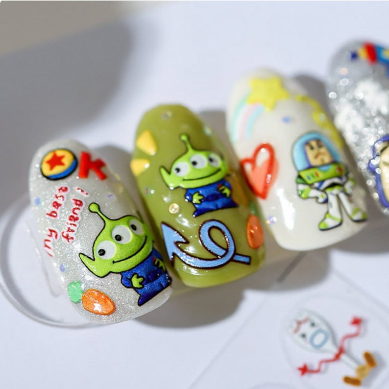 May include: Close-up of five fingernails with different designs. The designs include a green alien, a blue arrow, a heart, a cartoon astronaut, and a silver glitter design. The text "my best friend!" is written on one of the nails.