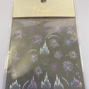 May include: Nail art stickers featuring a dark background with holographic fireworks and castle designs. The package includes the brand name "tomoni nailart" in gold lettering. The stickers also have Japanese text.