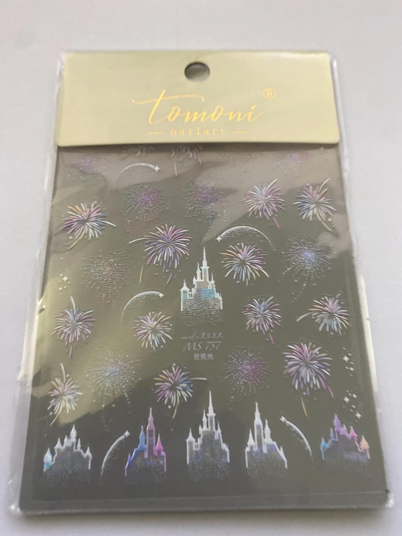 May include: Nail art stickers featuring a dark background with holographic fireworks and castle designs. The package includes the brand name "tomoni nailart" in gold lettering. The stickers also have Japanese text.