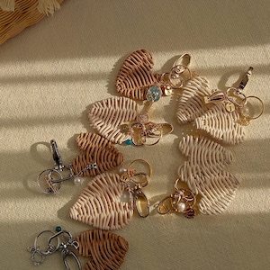 Op de afbeelding: Een verzameling hartvormige sleutelhangers gemaakt van geweven riet in beige en bruine tinten. Elke sleutelhanger heeft een metalen sluiting en decoratieve bedels, waaronder parels en kleine edelstenen. De sleutelhangers zijn gerangschikt op een neutrale ondergrond.