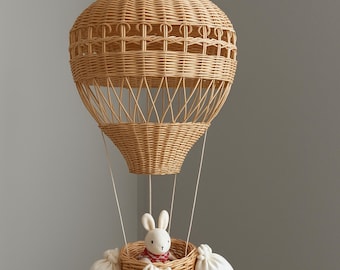 Op de afbeelding: Een hanglamp in de vorm van een luchtballon, gemaakt van gevlochten riet. De lamp heeft een ronde bovenkant en een mandje eronder, met een knuffelkonijn met een rood-witte sjaal. Witte zakken hangen aan de mand.