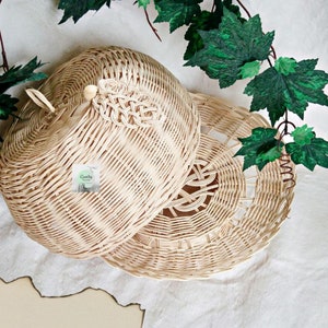 May include: A woven wicker dome-shaped food cover with a handle. The cover is made of natural light brown wicker and has a woven design. The cover is open and the base is visible. The base is a round woven wicker plate with a decorative woven design.