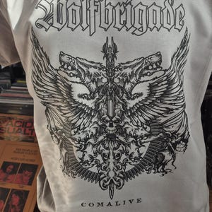 Puede incluir: Camiseta gris con el nombre de la banda "Wolfbrigade" en letras góticas. Debajo, un gráfico negro detallado de dos cabezas de lobo, alas, una calavera y una espada. La palabra "COMALIVE" está impresa debajo.