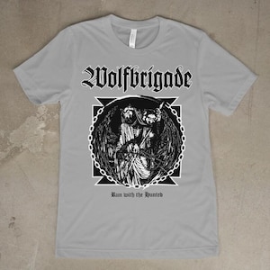 Könnte beinhalten: Ein hellgraues T-Shirt mit dem Bandnamen "Wolfbrigade" in gotischer Schrift über einer schwarz-weißen Grafik. Die Grafik zeigt eine gefesselte Figur mit Flügeln. Der Text "Run with the Hunted" steht darunter.