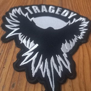 May include: Black and white embroidered patch with a stylized bird design and the word "TRAGEDY" in white lettering.