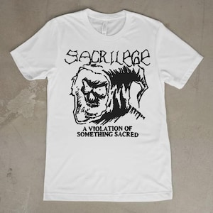 Può includere: Una t-shirt bianca a maniche corte con una stampa grafica nera sul petto. Il design include la parola SACRILEGE in un carattere stilizzato sopra un'illustrazione di un teschio incappucciato, con il testo A VIOLATION OF SOMETHING SACRED.