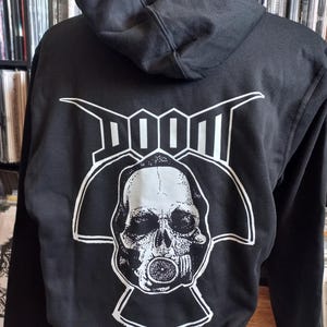 May include: Black hooded sweatshirt with a white graphic on the back. The graphic features the word "DOOM" above a detailed skull wearing a respirator. The sweatshirt has a drawstring hood and long sleeves.