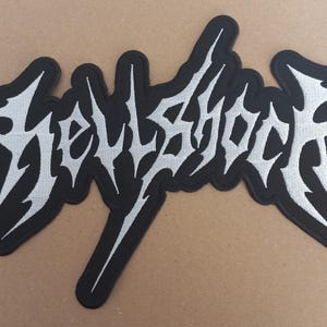 May include: Black and white embroidered patch with the word "HELLSHOCK" in a stylized font.