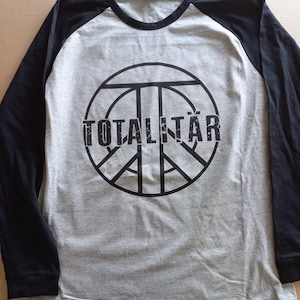 Op de afbeelding: Een grijs en zwart longsleeve shirt met een peace-teken graphic en het woord "TOTALITÄR" gedrukt op de voorkant.