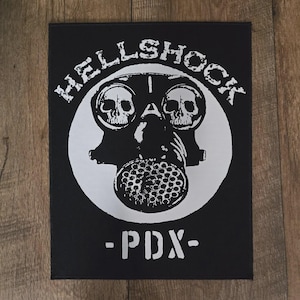 Könnte beinhalten: Ein rechteckiger schwarzer Stoffaufnäher mit einem weißen Siebdruckmotiv einer Gasmaske mit Totenköpfen in den Augenlinsen. Der Schriftzug HELLSHOCK ist über der Maske gebogen und der Text - PDX - ist in Weiß unter dem runden Motiv gedruckt.