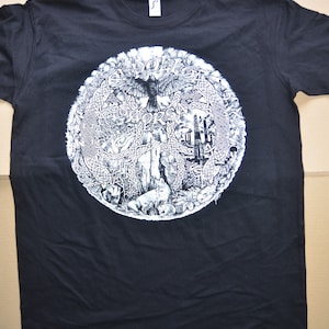 Op de afbeelding: Zwart T-shirt met een wit cirkelvormig grafisch ontwerp met een vogel, bomen en andere natuur-geïnspireerde elementen. De grafiek is omgeven door een cirkelvormige rand met een Keltisch knoopontwerp.