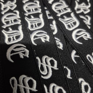May include: Close-up of several black fabric patches embroidered with the word "CRUEL" in white gothic lettering. The patches are stacked, showing the detailed stitching and texture of the fabric.