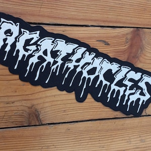 Pode incluir: Patch preto e branco com a palavra "RESCUE" em uma fonte gotejante. O patch é retangular com cantos arredondados e está colocado em uma superfície de madeira.