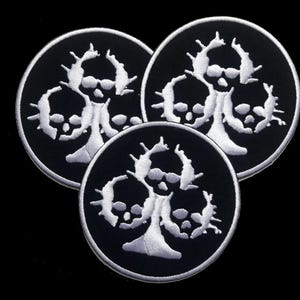 Puede incluir: Tres parches bordados en blanco y negro con un diseño de calavera. Las calaveras están dispuestas en forma de trébol.