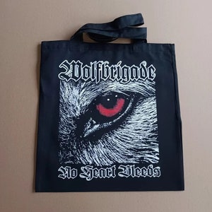 Può includere: Borsa tote nera con un occhio di lupo bianco e un'iride rossa. Il testo "Wolfrigade" e "No Heart Bleeds" è stampato in bianco sulla borsa.