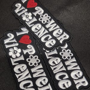 Può includere: Toppe nere ricamate con le parole "I Power Violence" in bianco, con un cuore rosso e decorazioni floreali. Le toppe sono rettangolari e hanno un bordo nero.