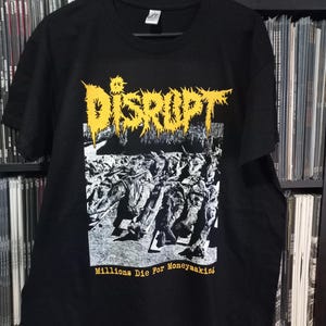 May include: Black t-shirt with a yellow graphic print of the word "DISRUPT" in a stylized font. The graphic also includes a black and white image of a crowd of people. The text "Millions Die For Moneymaking" is printed in yellow below the graphic.