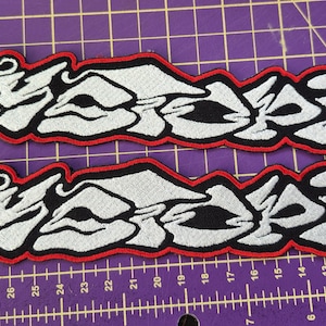 May include: Two embroidered patches with the word "ASSAIL" in a stylized, graffiti-style font. The text is white with black accents and a red border. The patches are displayed on a purple cutting mat.