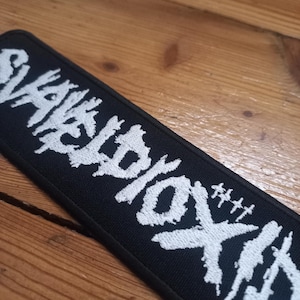 Peut inclure: Écusson brodé noir avec le texte blanc "SLAYER" dans une police stylisée.