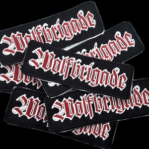 Peut inclure: Plusieurs écussons rectangulaires noirs avec le mot "Wolfbrigade" brodé en police gothique. Le texte est rouge avec un contour blanc. Les écussons sont superposés.