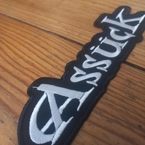 Könnte beinhalten: Schwarzer Patch mit weiß gesticktem "Assück"-Text.