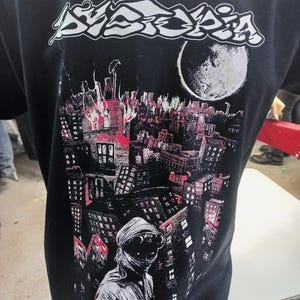 Może przedstawiać: Czarny T-shirt z białym nadrukiem nazwy zespołu "Dyspepsia" i panoramą miasta z dużym księżycem. Na dole koszulki znajduje się napis "EUROPEAN TOUR 2000".
