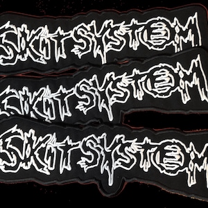 Può includere: Tre toppe nere ricamate con la scritta bianca "SKIT SYSTEM". Le toppe, di forma rettangolare curva, sono progettate per essere cucite su abbigliamento o accessori. L'estetica generale evoca la cultura punk o alternativa.