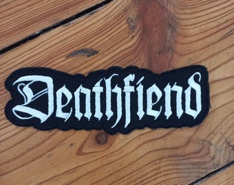 Peut inclure: Un écusson noir avec le texte blanc "Deathfiend" en police gothique. L'écusson est rectangulaire avec un bord ondulé. Le fond est une surface en bois avec le grain visible.