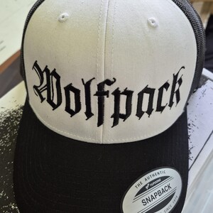 Może przedstawiać: Czarno-biała czapka typu trucker z napisem "Wolfpack" wyhaftowanym gotycką czcionką z przodu. Czapka ma czarny daszek i siatkową tylną część, z białym panelem przednim. Na daszku znajduje się naklejka "Snapback".