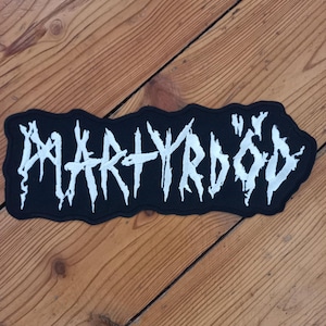 May include: Black and white patch with the word "MARTYRDÖD" in a jagged, gothic font. The patch has a black border and is rectangular in shape. The patch is laying on a wooden surface.