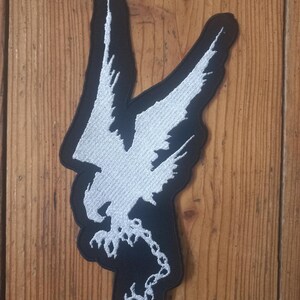 Op de afbeelding: Wit geborduurde patch van een vogel met gespreide vleugels, die een ketting in zijn klauwen vasthoudt, op een zwarte achtergrond.