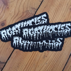 Puede incluir: Tres parches negros con la palabra "AGATHOCLES" en blanco, con fuente goteante. Los parches son rectangulares con un borde negro y están dispuestos sobre una superficie de madera. Estos parches son probablemente para ropa o accesorios.
