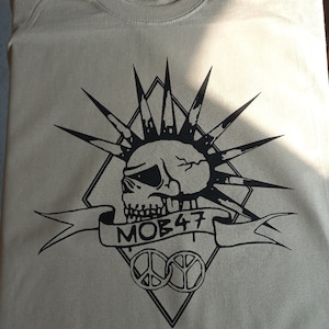 May include: A khaki t-shirt with a black graphic design of a skull with a mohawk, a banner that says "MOB 47", and two peace signs.