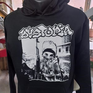 Puede incluir: Sudadera con capucha negra con un estampado gráfico blanco de una persona sosteniendo un rifle y la palabra "Dystopia" en letras de estilo graffiti.