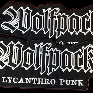Könnte beinhalten: Zwei schwarze gestickte Patches mit weißen Nähten. Der obere Patch sagt "Wolfpack" in einer gotischen Schrift. Der untere Patch sagt "Wolfpack" in der gleichen Schrift und "Lycanthro Punk" in einer kleineren Schrift darunter.