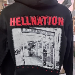Könnte beinhalten: Schwarzer Kapuzenpullover mit einem roten Grafikdruck, der "HELLNATION" und "OBEDIENCE TO THE LAW IS FREEDOM" sagt, mit einem Schwarzweißbild eines Gefängnishofs mit einem Zaun und einer Person, die im Hintergrund steht.
