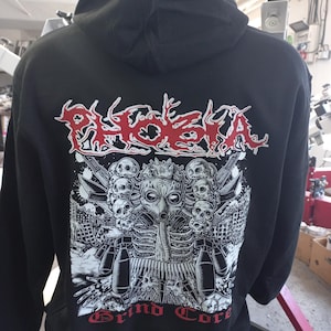May include: Black hooded sweatshirt with a white and black graphic print of a skull wearing a gas mask and surrounded by other skulls and weapons. The text "PHOEBA Grind Core" is printed in red below the graphic.