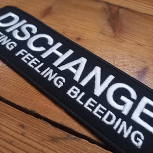 May include: Black embroidered patch with white text that reads "DISCHARGE" and "ING FEELING BLEEDING".